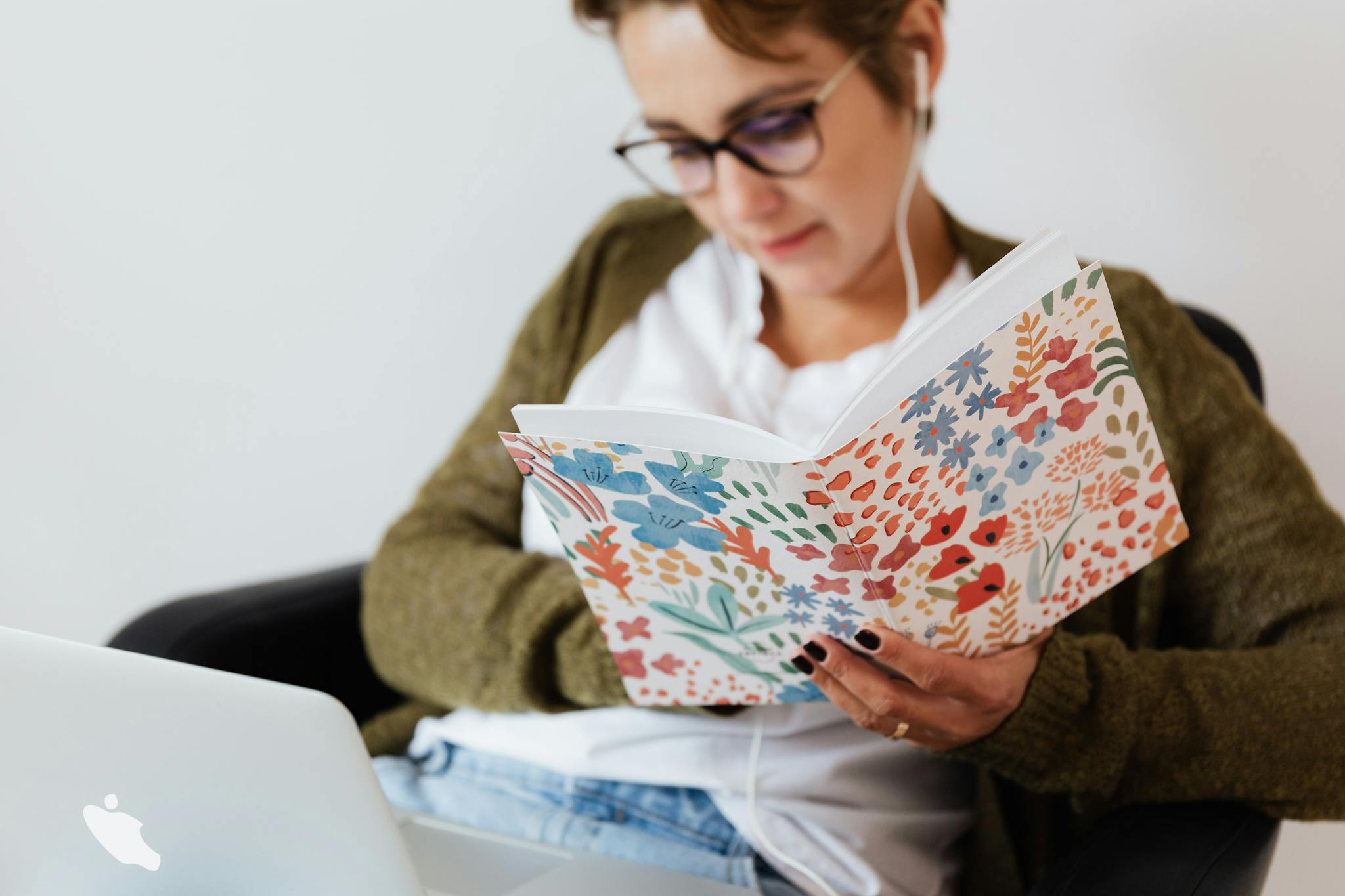 A woman reading a floral notebook while listening to music on her laptop indoors.