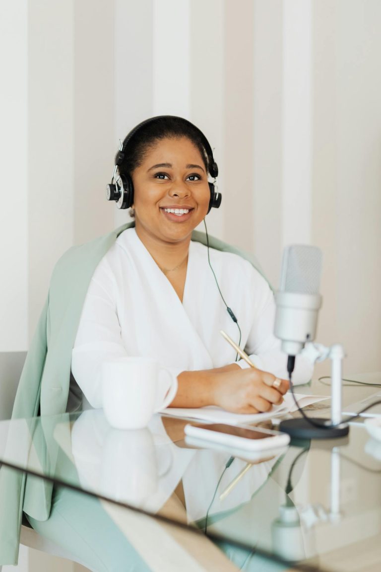 Smiling woman with headphones recording a podcast at home office with a microphone and tablet.
