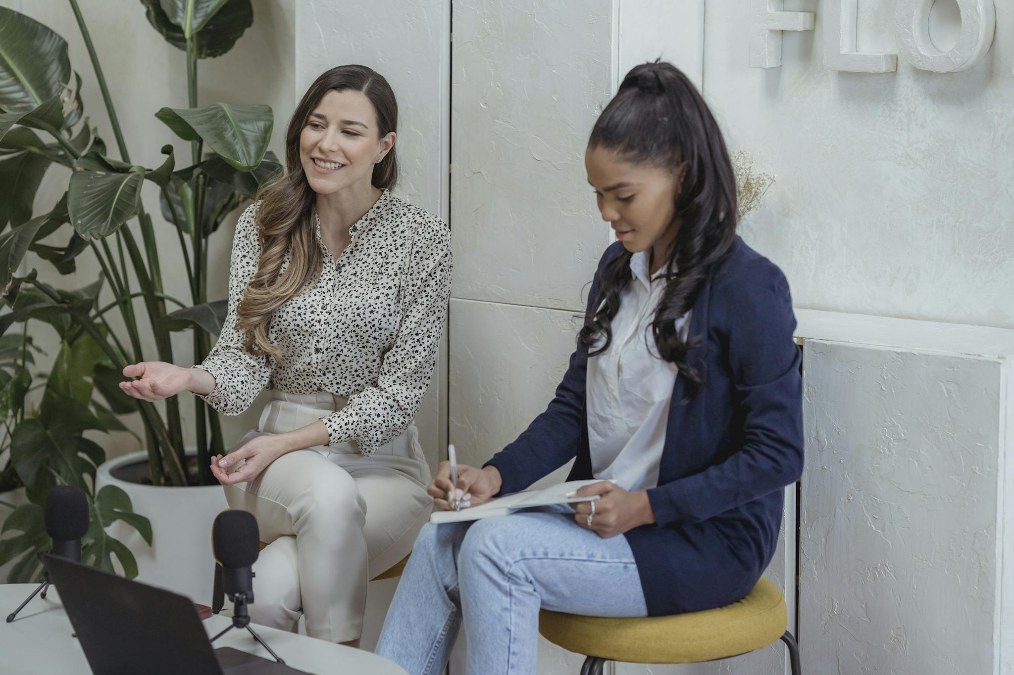 Two women engaged in a podcast interview setting, discussing and writing indoors.